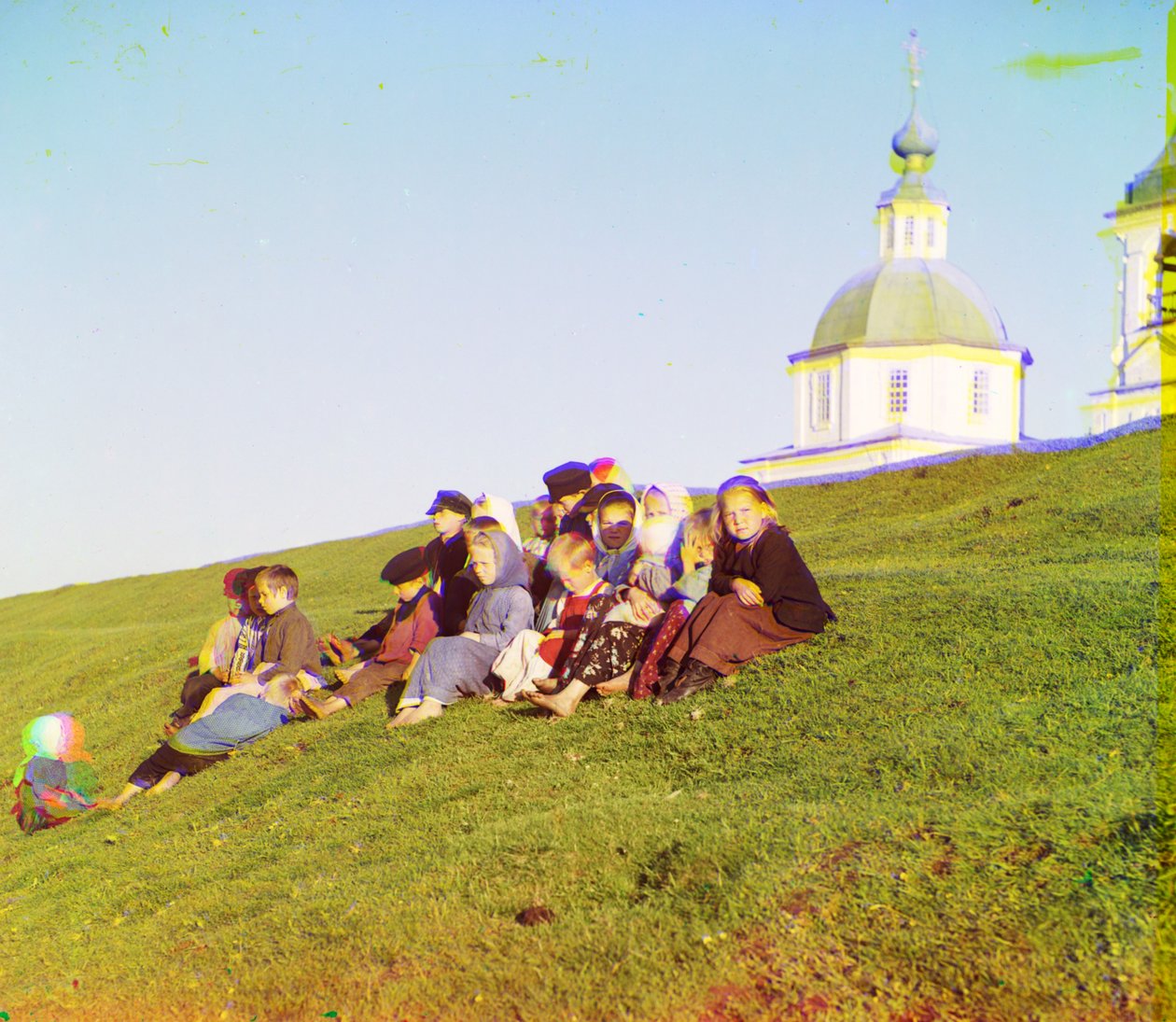 Russische Kinder sitzen auf einem Hügel in der Nähe einer Kirche und eines Glockenturms auf dem Land in der Nähe des Weißen Sees, im Norden des europäischen Russlands, 1905-1915 von Sergey Prokudin Gorsky